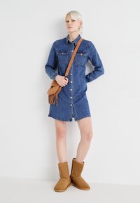 Denimklänning med långa ärmar, knappstängning, spetsig krage, två bröstfickor. Kombineras med bruna mockaankelboots och en tan crossover-väska.