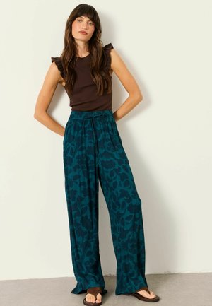 Woman with long brown hair wearing a sleeveless dark brown top and loose teal floral pants, standing with hands behind back.