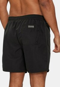 Pantaloni da bagno neri con vita elastica, tasca posteriore e patch con logo "GUESS" sulla tasca. Tessuto morbido.