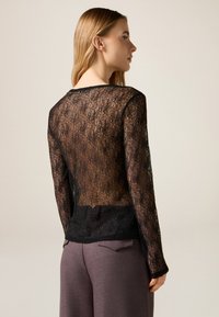 Top de encaje negro con patrón floral, mangas largas y escote redondeado, superpuesto sobre una tela negra sólida en la parte inferior.