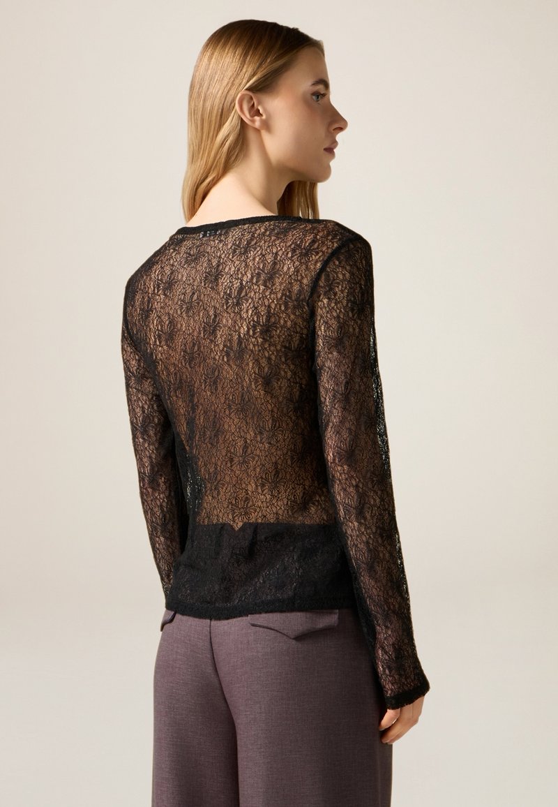 Top de encaje negro con patrón floral, mangas largas y escote redondeado, superpuesto sobre una tela negra sólida en la parte inferior.