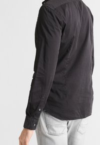 Camicia nera a maniche lunghe con colletto abbottonato, texture liscia e design aderente. Presenta polsini abbottonati e orlo curvato.