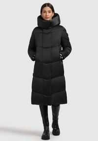 Schwarzer langer Steppmantel mit hohem Kragen, der gesteppte Abschnitte, Seitentaschen und glattes Gewebe aufweist. Dazu werden kniehohe schwarze Stiefel getragen.