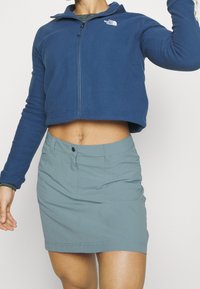 Osoba v modrém zkráceném fleece od North Face a světlém modrém těsně přiléhajícím sukni, ukazující břicho a část zvednuté paže.