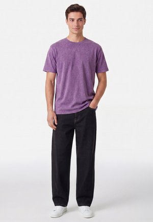 Junger Mann steht, trägt ein gewaschenes lila T-Shirt, lockere schwarze Jeans und weiße Sneaker mit einer Hand in der Tasche vor einem einfarbigen Hintergrund.