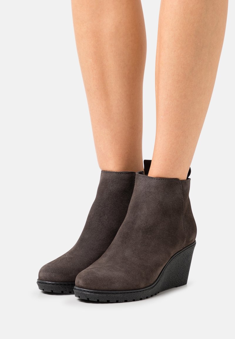 Bottes compensées en suède gris foncé montant à la cheville, portées sur des jambes nues, avec des semelles noires texturées et une languette à l'arrière.