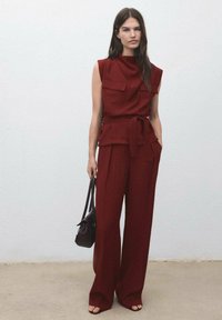 Burgundský dvoudílný outfit s bezrukávovým topem, který má dvě kapsy na prsou, a odpovídajícími širokými kalhotami, které jsou stahovány v pase. Hnědá kabelka.