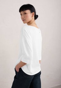 Bílý, lehký top s volným střihem, 3/4 rukávy, kulatý výstřih na zádech a strukturovaný povrch. V kombinaci s tmavými džínovými kalhotami.