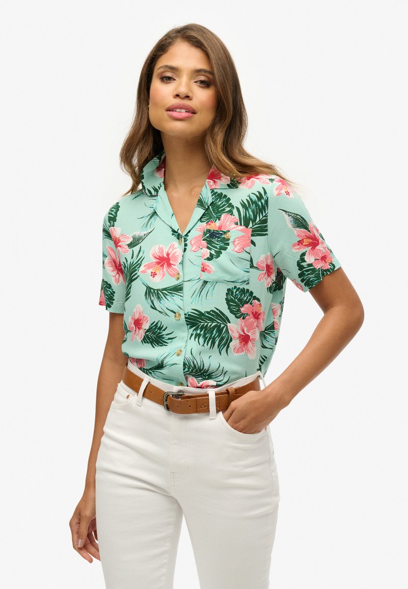 Camicia a maniche corte con colletto in verde menta con motivi floreali rosa e foglie verdi; dotata di taschino sul petto e chiusura con bottoni sul davanti.