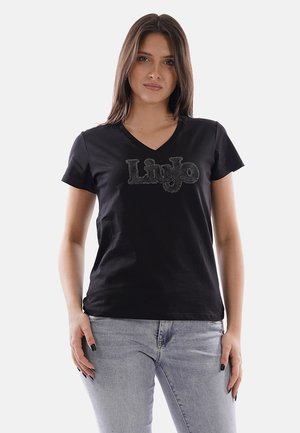 Donna con capelli castani lisci che indossa una maglietta nera a scollo a V con logo "Liu Jo" in strass e jeans grigio chiaro, in piedi davanti a uno sfondo bianco.