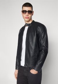 Chaqueta de cuero negra con un cuello alto, cierre de cremallera y bolsillos laterales. Llevada sobre una camiseta blanca, combinada con jeans oscuros y gafas de sol.