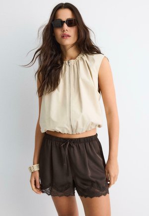 Femme aux longs cheveux bruns portant des lunettes de soleil noires, un haut beige sans manches avec fermeture éclair, un short marron foncé à cordon avec bordure en dentelle, et un bracelet beige.