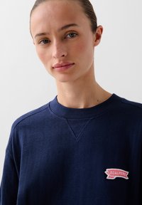 Felpa blu navy con collo a giro, tessuto texturizzato e un patch con il logo "SCALPERS" rosso e beige sul lato sinistro del petto.