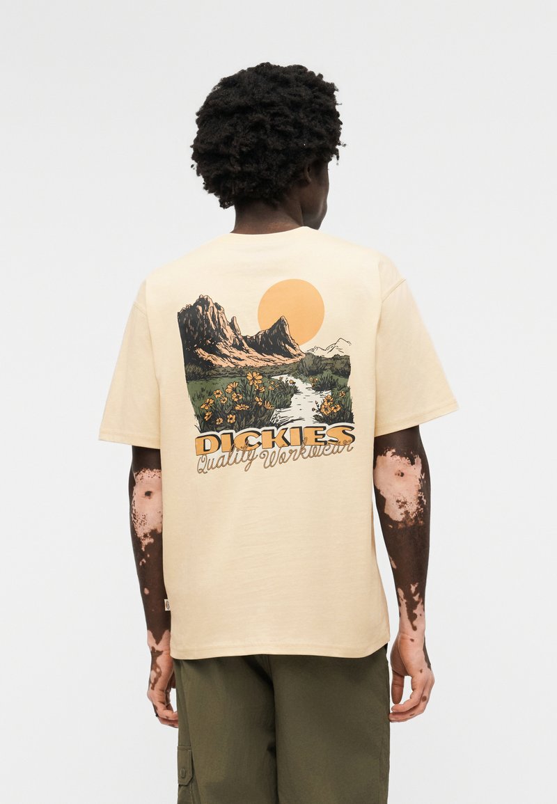Person mit dunkler Haut und Vitiligo, die ein beige T-Shirt trägt, auf dem ein Berg, ein Fluss, die Sonne und Blumen mit dem Schriftzug "Dickies Quality Workwear" auf dem Rücken zu sehen sind.