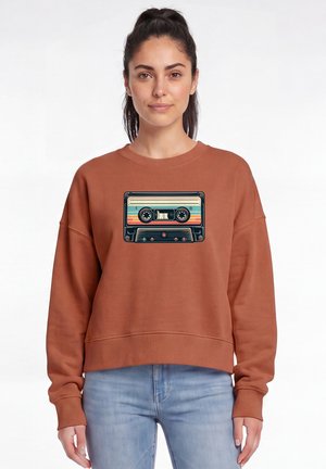 Frau mit dunklem Haar im Pferdeschwanz trägt einen rostfarbenen Sweatshirt mit einer bunten Retro-Kassettengrafik und hellblauen Jeans.