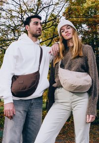 Två individer som bär crossbody-väskor står utomhus. Den ena väskan är brun, den andra är ljusbeige, båda har släta tyger och enkla mönster.