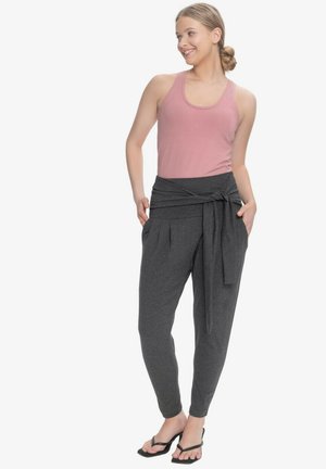 Uśmiechnięta kobieta z włosami związanymi do tyłu, ubrana w różowy top na ramiączkach, ciemnoszare spodnie z wysokim stanem i paskiem w talii oraz czarne sandały na obcasie, ręce w kieszeniach.