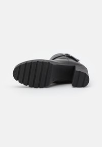 Černá kožená bota s masivní černou podrážkou, s vodorovnými drážkami pro větší přilnavost a dekorativní sponou na boku.