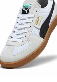Sneaker bianca in mesh e suede con dettagli neri. Presenta lacci bianchi, suola in gomma e un logo PUMA verde sulla linguetta.