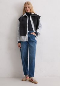 Czarna bezrękawnik na paskowanej koszuli zapinanej na guziki, w połączeniu z niebieskimi dżinsami i beżowymi trampkami z czerwonymi akcentami. Płaskie, teksturowane tło.