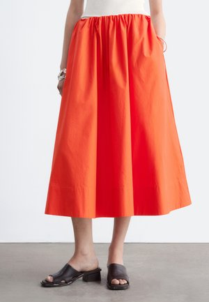 Middenlange feloranje rok gedragen met zwarte open teen blokhak sandalen door een persoon die op een betonnen vloer staat.
