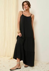 Robe maxi noire avec des bretelles fines, au design en couches et en tissu doux. Associée à des sandales plates, présentant un design tressé.