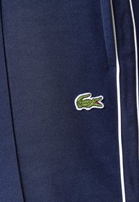 Marineblå joggebukser med hvite sidestriper og brodert grønn krokodillelogo. Teksturert overflate, tettsittende design og minimalistisk stil.