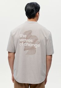 Šedé bavlněné tričko s krátkým rukávem, které na zadní straně obsahuje velkou grafiku s textem "vlny změny" a abstraktním designem.