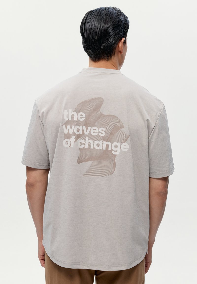 Šedé bavln�ěné tričko s krátkým rukávem, které na zadní straně obsahuje velkou grafiku s textem "vlny změny" a abstraktním designem.