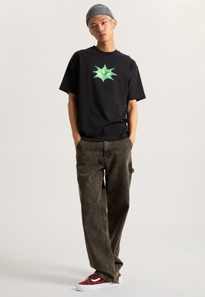 Zwart T-shirt met een groen grafisch ontwerp, gecombineerd met losse bruine cargobroeken. Model draagt rode en witte sneakers. Neutrale achtergrond.