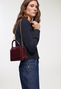 Piccola borsa a mano in pelle bordeaux con finitura lucida, forma strutturata, manico superiore e tracolla a catena, presentata su uno sfondo chiaro.
