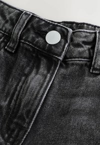 Läheltä kuvattu musta denimkangas, joka esittelee korkeavyötäröisen muotoilun, hopeisen napin ja kontrastivärisen ompelun vyötäröllä.