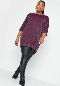 Burgundzka tunika z krótkimi rękawami i bocznymi rozcięciami, w połączeniu z błyszczącymi czarnymi leginsami z sztucznej skóry oraz dopasowanymi botkami.