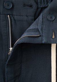 Gros plan d’un pantalon bleu marine montrant une fermeture éclair argentée partiellement ouverte, un bouton de fermeture et un cordon beige à l’intérieur de la taille.