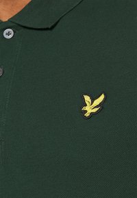 Dunkelgrünes Poloshirt aus strukturiertem Gewebe mit einer Zwei-Knopf-Leiste. Auf der linken Brust befindet sich ein gelbes, gesticktes Vogel-Logo.