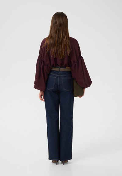 Blouse bordeaux à manches larges et volantes, associée à un pantalon en denim foncé taille haute. Un sac vert est tenu dans la main gauche.