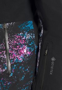 Gros plan sur une veste noire avec un motif tacheté coloré bleu et rose, deux fermetures éclair verticales étanches et logo GORE-TEX sur la manche.