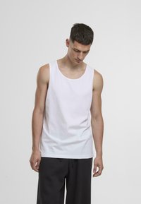 Fehér pamut tank top, kerek nyakkal, ujjatlan kivitelben és laza szabásban. Sötét nadrággal viselve egy egyszínű háttér előtt.