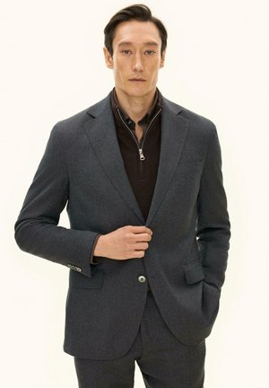 Mann trägt eine dunkelgraue Anzugsjacke über einem hochgeschlossenen dunklen Hemd, mit einer Hand richtet er die Jacke und die andere ist in der Tasche.