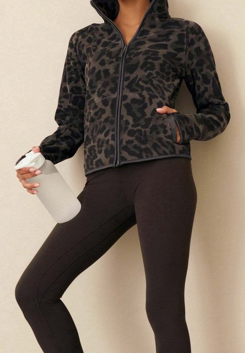 Personne portant une veste zippée noir et gris à imprimé léopard et des leggings noirs, tenant une bouteille d'eau en plastique transparent dans une main.