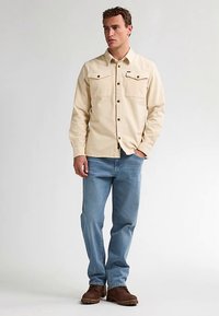 Beige pitkähihaisen paidan edessä kaksi rintataskua ja napitus, yhdistetty vaaleansinisiin farkkuihin ja ruskeisiin kenkiin, seisoo neutraalia taustaa vasten.