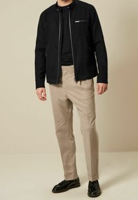 Schwarze leichte Jacke mit durchgehendem Reißverschluss, Stehkragen und Brusttasche, kombiniert mit beigen maßgeschneiderten Hosen und schwarzen Schuhen.