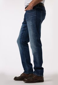 Vaqueros de mezclilla azul oscuro con un corte recto, que presentan un ligero desgaste y detalles desgastados. Combinados con zapatos de cordones marrón oscuro.