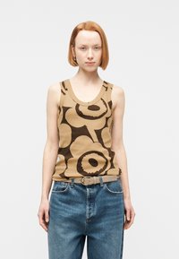 Νεαρό άτομο με κοντά κόκκινα μαλλιά που φοράει μπεζ αμάνικο τοπ με μεγάλα σκούρα καφέ αφηρημένα λουλούδια και φαρδιά μπλε τζιν με μπεζ ζώνη.