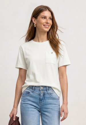 Ung kvinde med langt hår iført hvid kortærmet t-shirt og blå jeans, der holder en brun jakke i venstre hånd, smilende og kigger til siden.