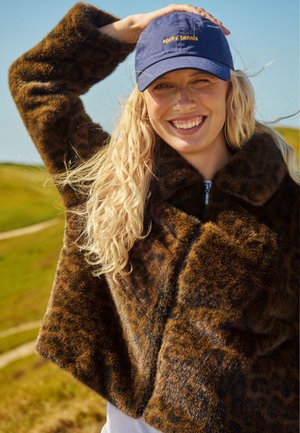 Femme souriante aux longs cheveux blonds porte un manteau en fourrure imprimé léopard et une casquette bleue en extérieur lors d'une journée ensoleillée.