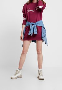 Burgundin värinen collegepuku, jossa on valkoiset raidat, sidottu farkkupaita vyötäröllä ja vaaleat, paksupohjaiset lenkkarit. Tasainen kangas.