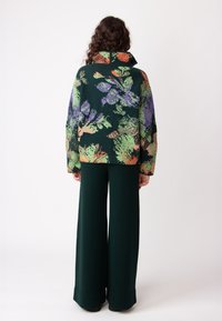 Pleteni jopič z visokim ovratnikom, ki prikazuje večbarvni cvetlični vzorec na temno zeleni podlagi; v kombinaciji z širokimi temno zelenimi hlačami.