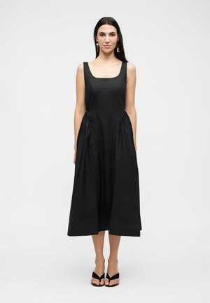 Γυναίκα με μακριά σκούρα μαλλιά, που φοράει μαύρο midi φόρεμα χωρίς μανίκια με τετράγωνο ντεκολτέ, πλαϊνές τσέπες, μαύρα πέδιλα ανοιχτής μύτης και κρεμαστά σκουλαρίκια.