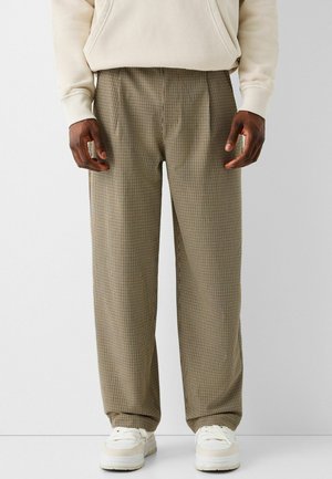 Homme portant un pantalon beige à carreaux, un sweat à capuche crème et des baskets blanches, debout devant un fond uni et clair.